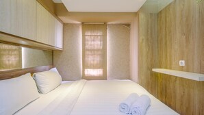 A neatly made bed with white linens and two rolled towels, a wooden headboard, and a wooden wall panel. - Modern 2BR at Bassura City Apartment (Jakarta)