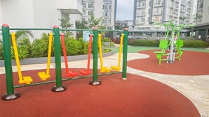 Un espace extérieur aménagé pour l’exercice, avec des balançoires et un appareil de musculation vert, entouré d’une surface rouge en caoutchouc et de verdure, avec des immeubles d’habitation en arrière-plan.