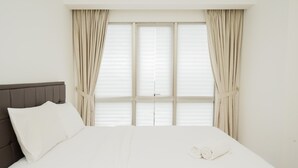 A neatly made bed with white linens and a folded towel, a grey upholstered headboard, and beige curtains on a window with blinds.