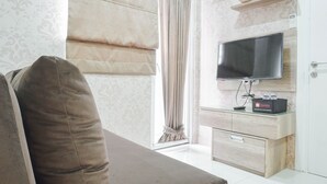 A modern living room with a sofa, a flat-screen TV mounted on a wooden cabinet, and a window with curtains. - Cozy Stay at 2BR Green Pramuka Apartment near Shopping Center (Jakarta)