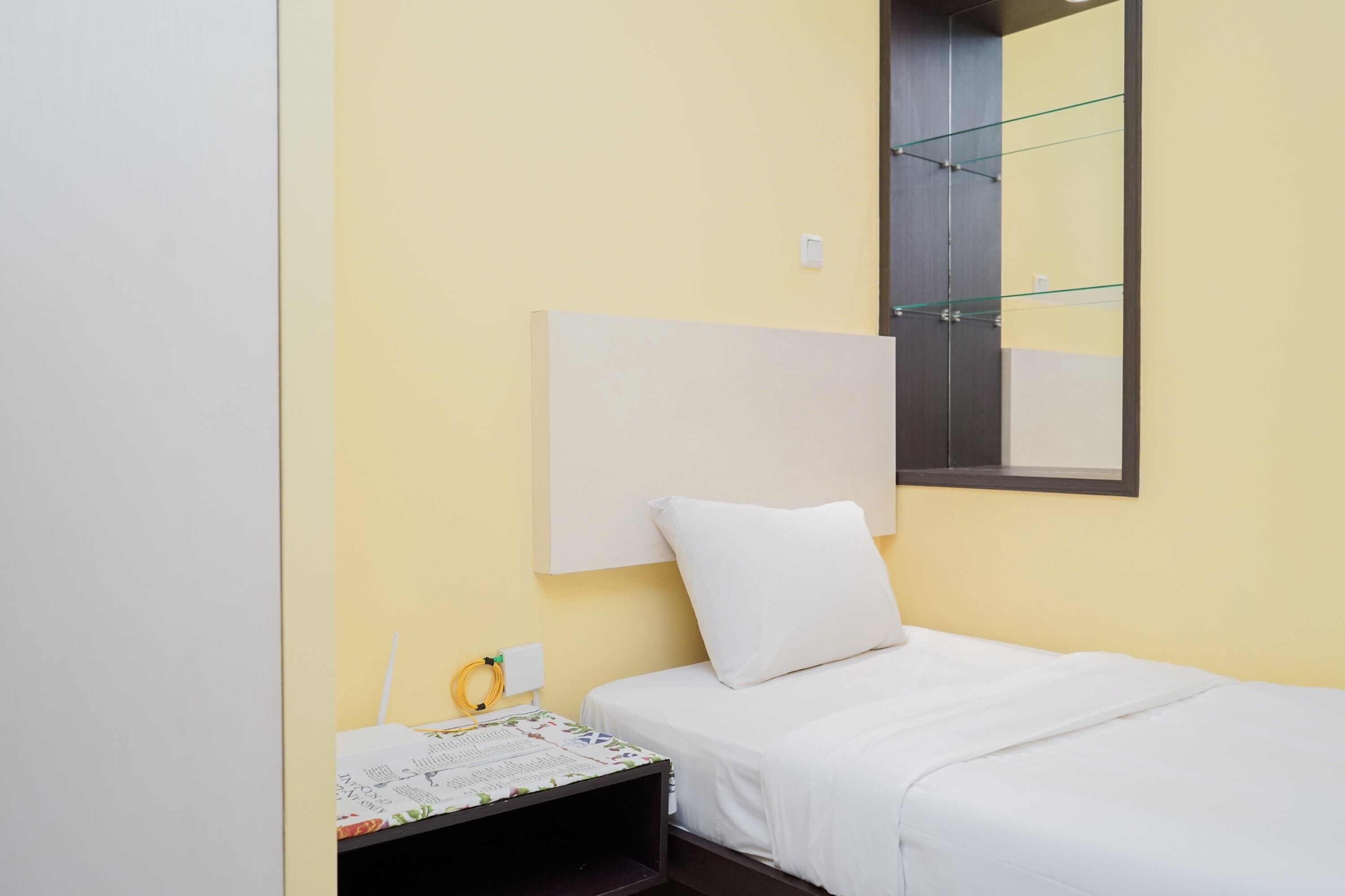 Una habitación pequeña y ordenada con una cama individual, una mesita de noche con lámpara, un espejo y una ventana.