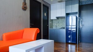 A modern living room with a white sofa and a bright orange chair, a kitchen with a tiled backsplash, and a wooden floor. - Exquisite & Spaciuos 1BR Apartment at Tamansari Papilio (Surabaya)