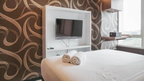 Ein Hotelzimmer mit einem an der weißen Wand befestigten Flachbildfernseher, einem Bett mit weißer Bettwäsche und einem Fenster mit Blick auf die Stadt.