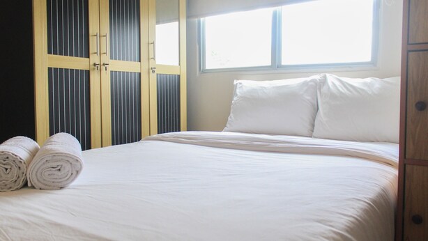 A neatly made bed with white linens and pillows, a window with blinds, and a wooden wardrobe with vertical slats. - Comfy and Best View 2BR at Serpong Greenview Apartment (South Tangerang)