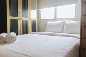 A neatly made bed with white linens and pillows, a window with blinds, and a wooden wardrobe with vertical slats. - Comfy and Best View 2BR at Serpong Greenview Apartment (South Tangerang)
