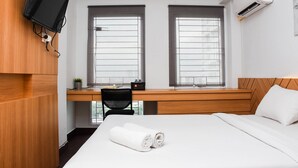 Habitación de hotel con escritorio de madera, dos ventanas con persianas, cama con ropa blanca, televisor montado en la pared y equipo de aire acondicionado.