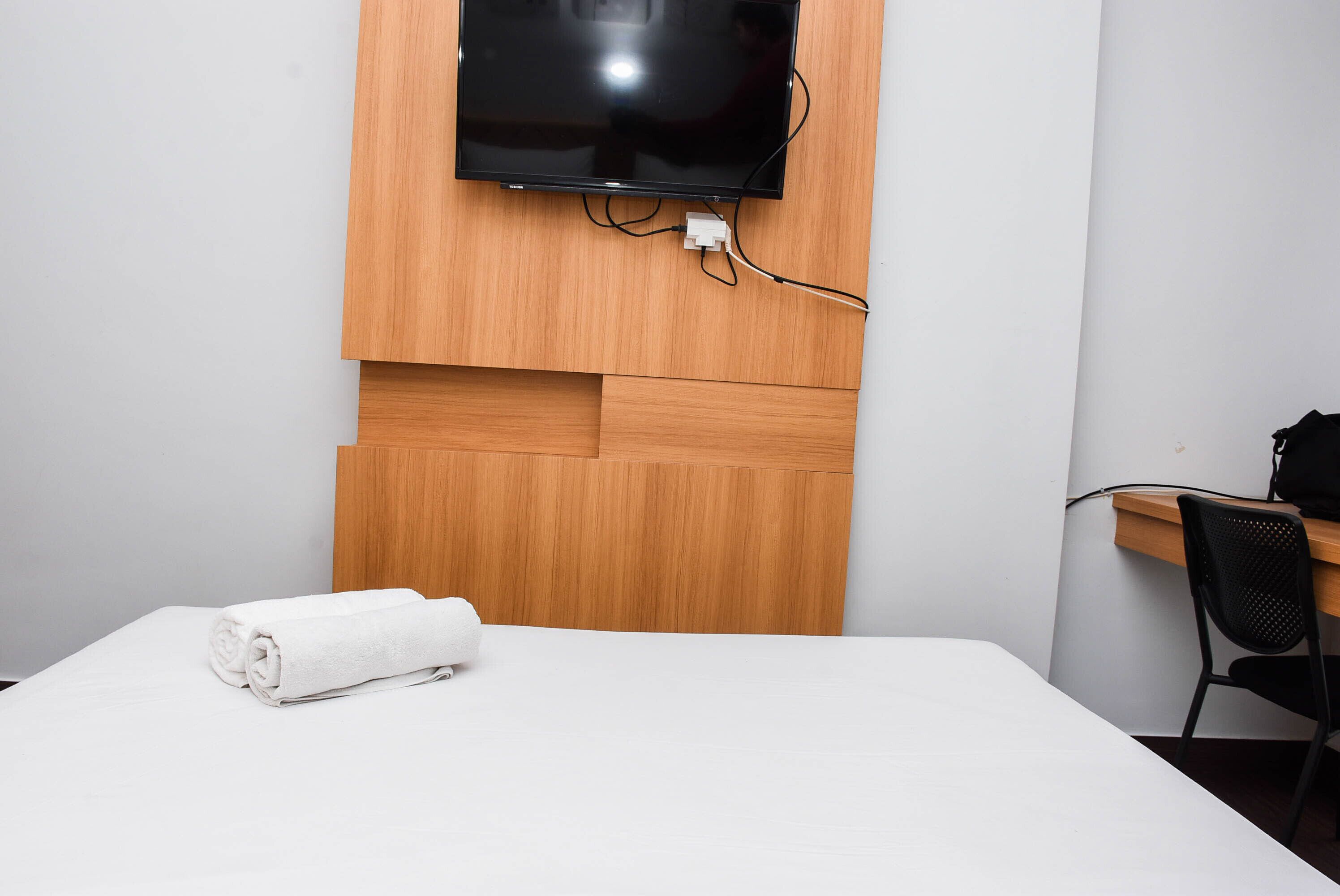 Une chambre d’hôtel avec une tête de lit en bois, un téléviseur à écran plat fixé au-dessus, un lit bien fait avec une serviette blanche, et un bureau avec une chaise.