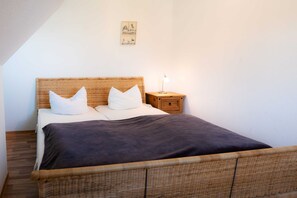 A bed with a dark blanket, a wicker headboard, and white pillows. There is a small wooden nightstand with a lamp beside the bed.