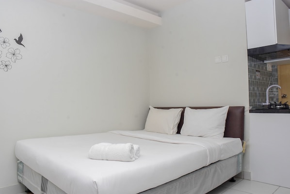 A neatly made bed with white linens and a folded towel, a brown headboard, and a small window with a view of the outside.