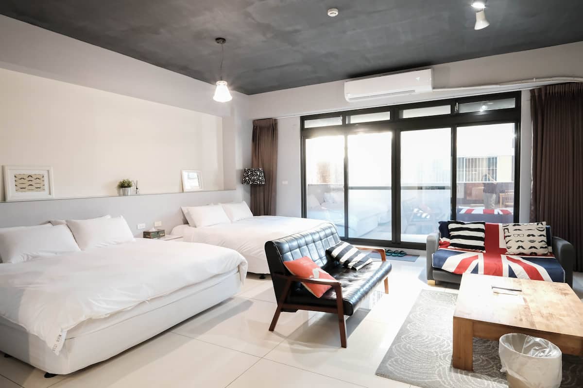 A modern hotel room with two beds, a sofa, a coffee table, and a view of the city through large windows.