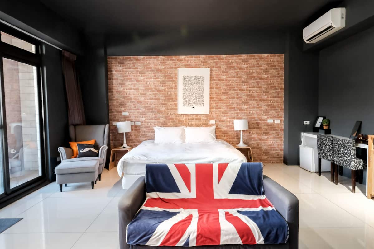 A modern hotel room with a large bed, a grey armchair, a small table with a lamp, and a view through a large window.