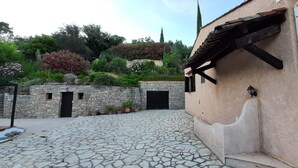Une cour pavée de pierres, bordée d’un mur en pierre, un bâtiment doté d’une porte noire et un jardin où poussent différentes espèces de plantes.