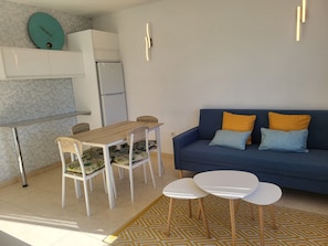 Un espace de vie compact comprenant un coin cuisine, une table à manger avec des chaises, un canapé avec des coussins et une petite table basse.