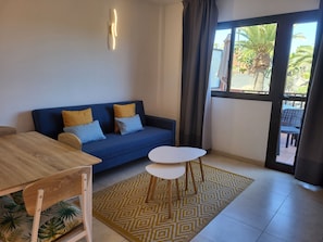 Une pièce avec un canapé bleu, une table en bois, une petite table blanche, un tapis à motifs et une vue sur des palmiers par la fenêtre.