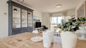 Un coin repas avec une table en bois, des objets décoratifs blancs, un meuble gris avec des étagères, un téléviseur et une vue sur l’extérieur.