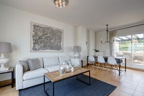 A modern living room with a sofa, coffee table, and dining area with chairs. - Penthouse Apartment in Alcaidesa with Mountain View, Wi-Fi, Pool, Garden & Terrace (Alcaidesa)