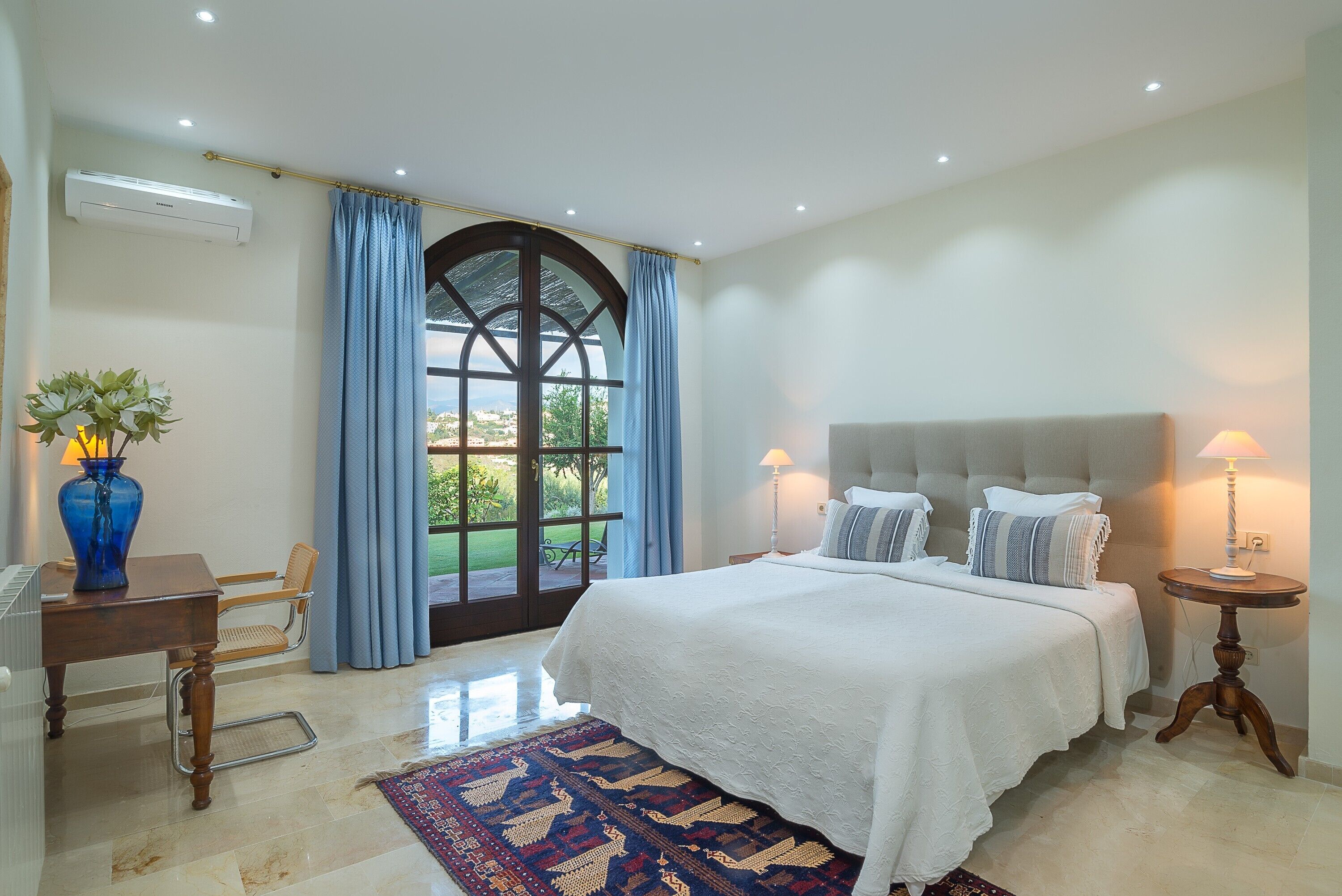 A bedroom with a large bed, a desk, a chair, a lamp, and a view through a window.