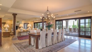 Un vaste espace dédié à la salle à manger, avec une grande table en bois, des chaises blanches, un lustre et une vue dégagée grâce à de grandes fenêtres.