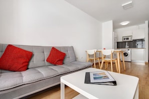 A modern living room with a grey sofa, red cushions, a white coffee table, and a kitchen area with a washing machine.