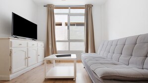 Een woonkamer met een flatscreen televisie op een witte kast, een grijze bank en een raam met gordijnen.