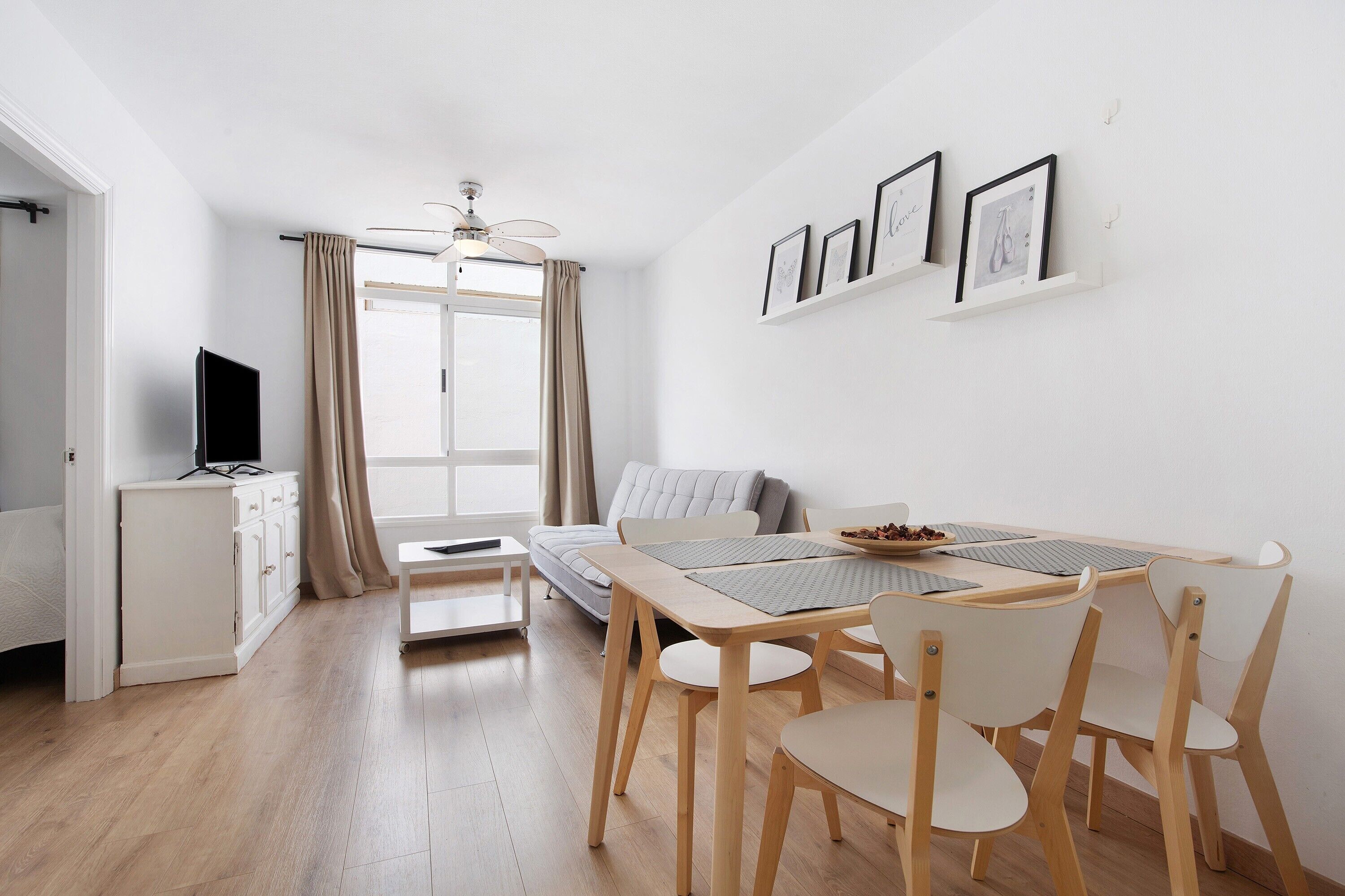 A modern living room with a sofa, a dining table with chairs, a TV on a cabinet, and a ceiling fan.