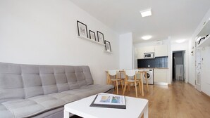 Een moderne woonkamer met een grijze bank, een witte salontafel en een eethoek met houten stoelen. In de achtergrond is een keuken met witte kasten en een magnetron zichtbaar.