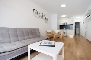 Un salon moderne avec un canapé gris, une table basse blanche et un coin repas avec des chaises en bois. On aperçoit au fond une cuisine avec des meubles blancs et un four à micro-ondes.
