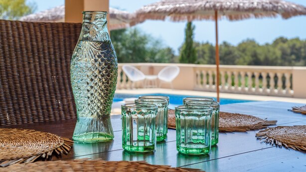 Una mesa con una botella de vidrio decorativa y varios vasos, frente a un fondo de muebles de mimbre y sombrillas, con vista a una zona de piscina.