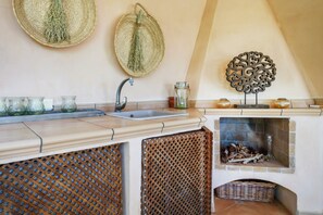 A kitchen with a sink, two hanging baskets with herbs, a decorative metal piece, and a fireplace with logs.