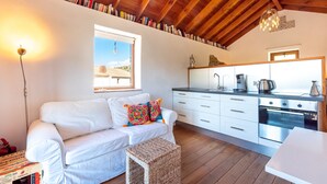 A cozy room with a white sofa, a small table, a kitchen area with white cabinets, and a wooden ceiling.