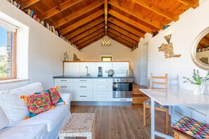 Um interior acolhedor com teto de madeira, sofá branco, almofadas coloridas, cozinha com eletrodomésticos modernos e uma área de jantar com mesa branca e cadeiras de madeira.
