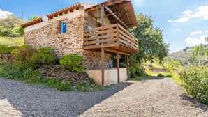 A stone house with a wooden balcony, surrounded by greenery and a gravel path.