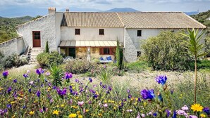 Une maison en pierre à deux étages, avec un toit en tuiles, un porche à l’avant et un jardin planté de fleurs sauvages.