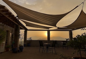 Un'area salotto esterna riparata con sedie, tavoli, piante in vaso e vista sul tramonto sulle montagne.