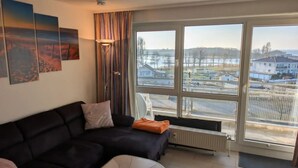 A living room with a black sofa, a radiator, and a large window with a view of a residential area. - Ostseepanorama Heiligenhafen – Apartment with Sea View (Heiligenhafen)