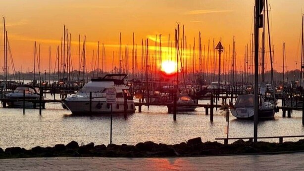 A marina with boats docked, a sunset in the background, and a paved walkway in the foreground. - Ostseepanorama Heiligenhafen – Apartment with Sea View (Heiligenhafen)