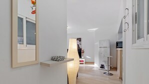 Ein schmaler Flur mit einem gerahmten Bild links, einer hohen Stehlampe in der Mitte und einem weißen Kühlschrank rechts.