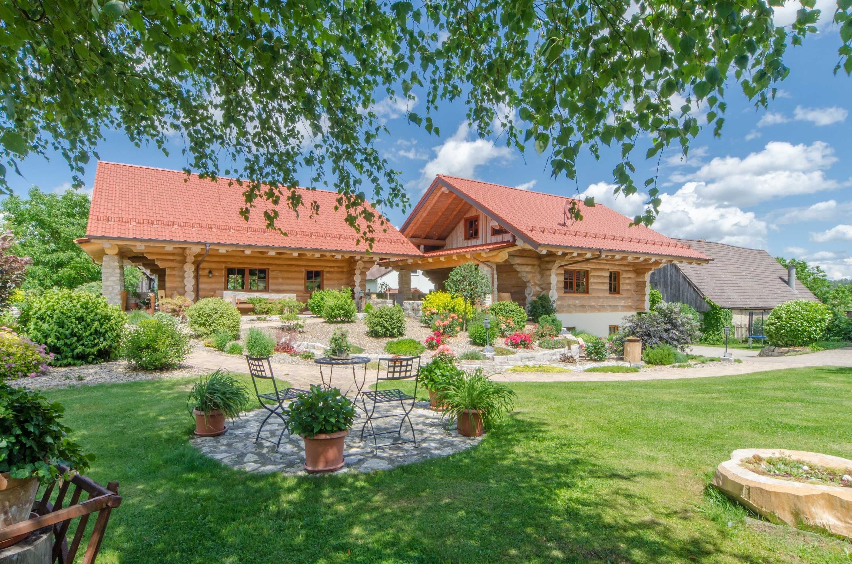 A two-story wooden house with a red roof, surrounded by a well-maintained garden with various plants and a patio area with outdoor furniture.