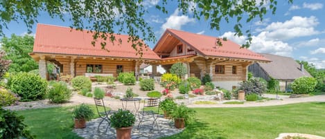 A two-story wooden house with a red roof, surrounded by a well-maintained garden with various plants and a patio area with outdoor furniture.
