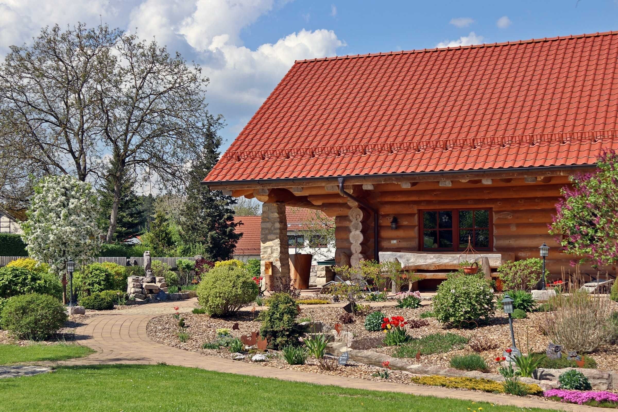 A log cabin with a red-tiled roof, surrounded by a landscaped garden with various plants and a winding path.