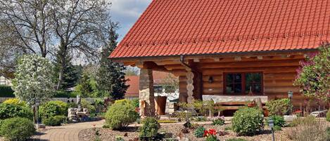 A log cabin with a red-tiled roof, surrounded by a landscaped garden with various plants and a winding path.