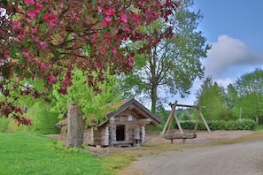 A rustic stone cabin with a wooden porch, surrounded by lush greenery and flowering trees.