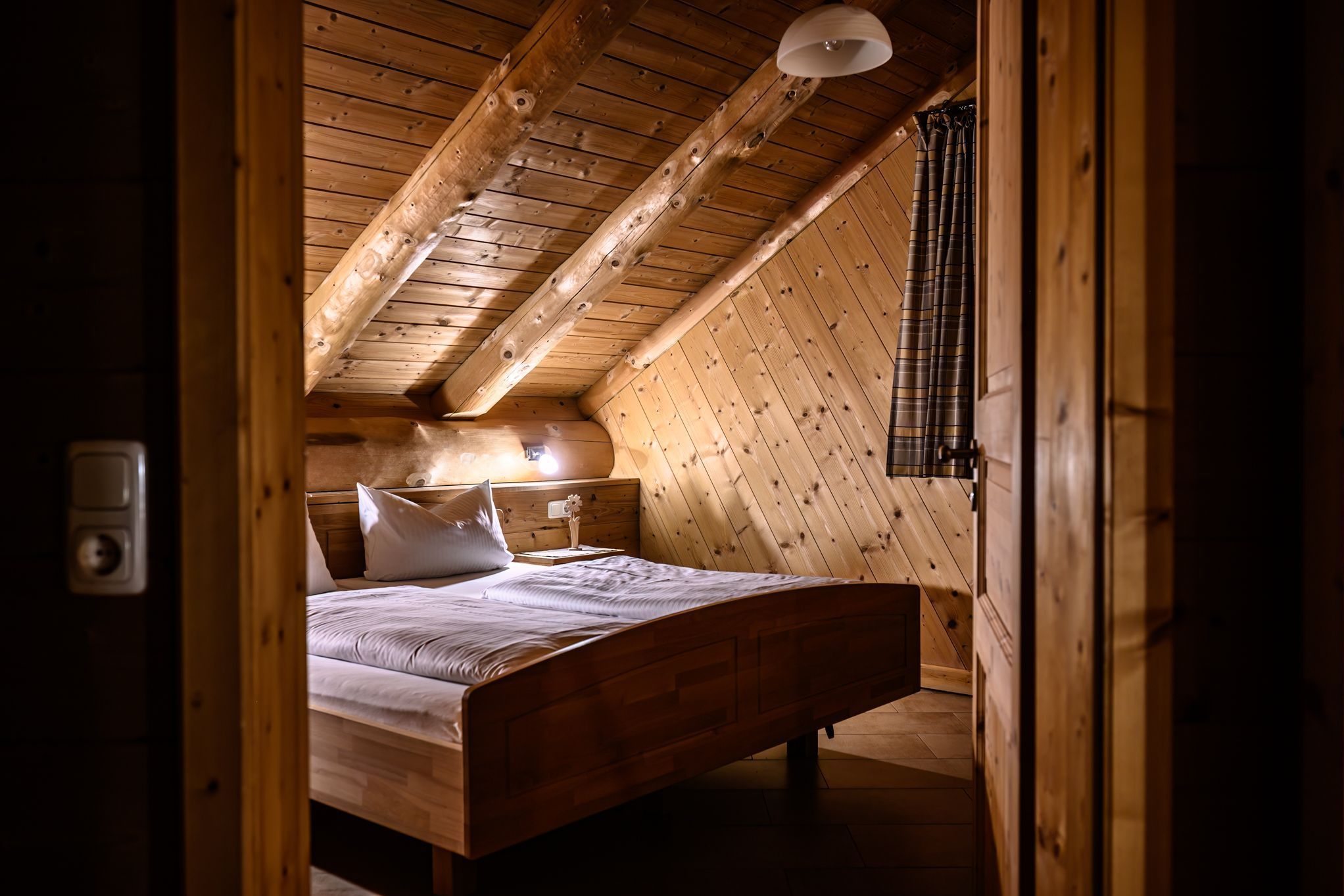 A wooden bedroom with a sloped ceiling, a bed, a bedside table, and a lamp.