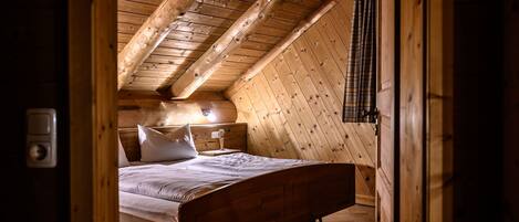 A wooden bedroom with a sloped ceiling, a bed, a bedside table, and a lamp.