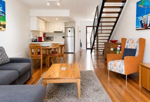 A modern living room with a sofa, coffee table, and patterned armchair. A kitchen with white cabinets and a stainless steel refrigerator is visible in the background.