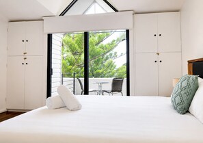 A bedroom with a large bed, a balcony with a table and chairs, and a view of trees.