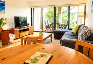 A modern living room with a TV, wooden furniture, a balcony with outdoor seating, and a view of greenery.
