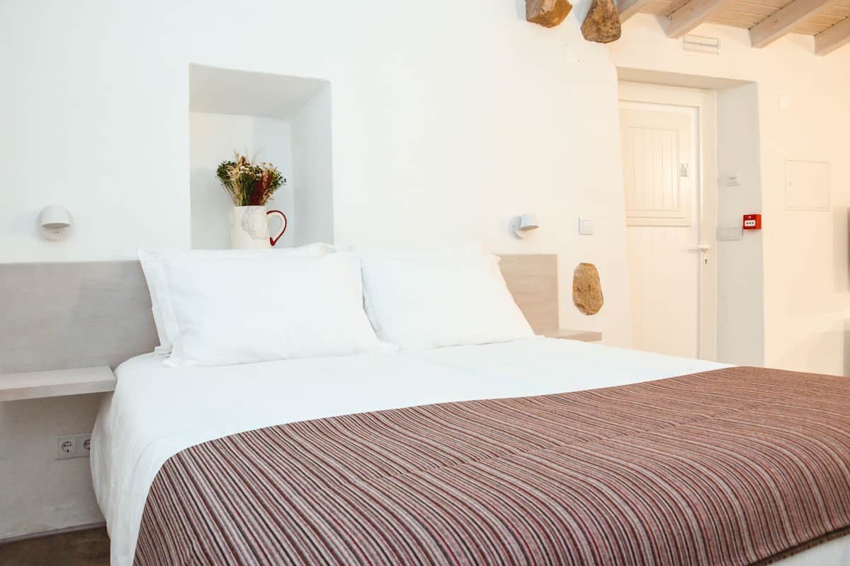 A neatly made bed with white linens and a striped blanket, a small vase with dried flowers, and a white wall with a wooden shelf.