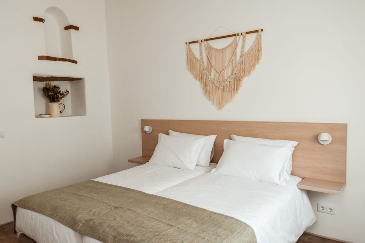 A neatly made bed with white linens and a beige throw blanket. There is a wooden headboard and a wall-mounted macrame decoration above the bed.