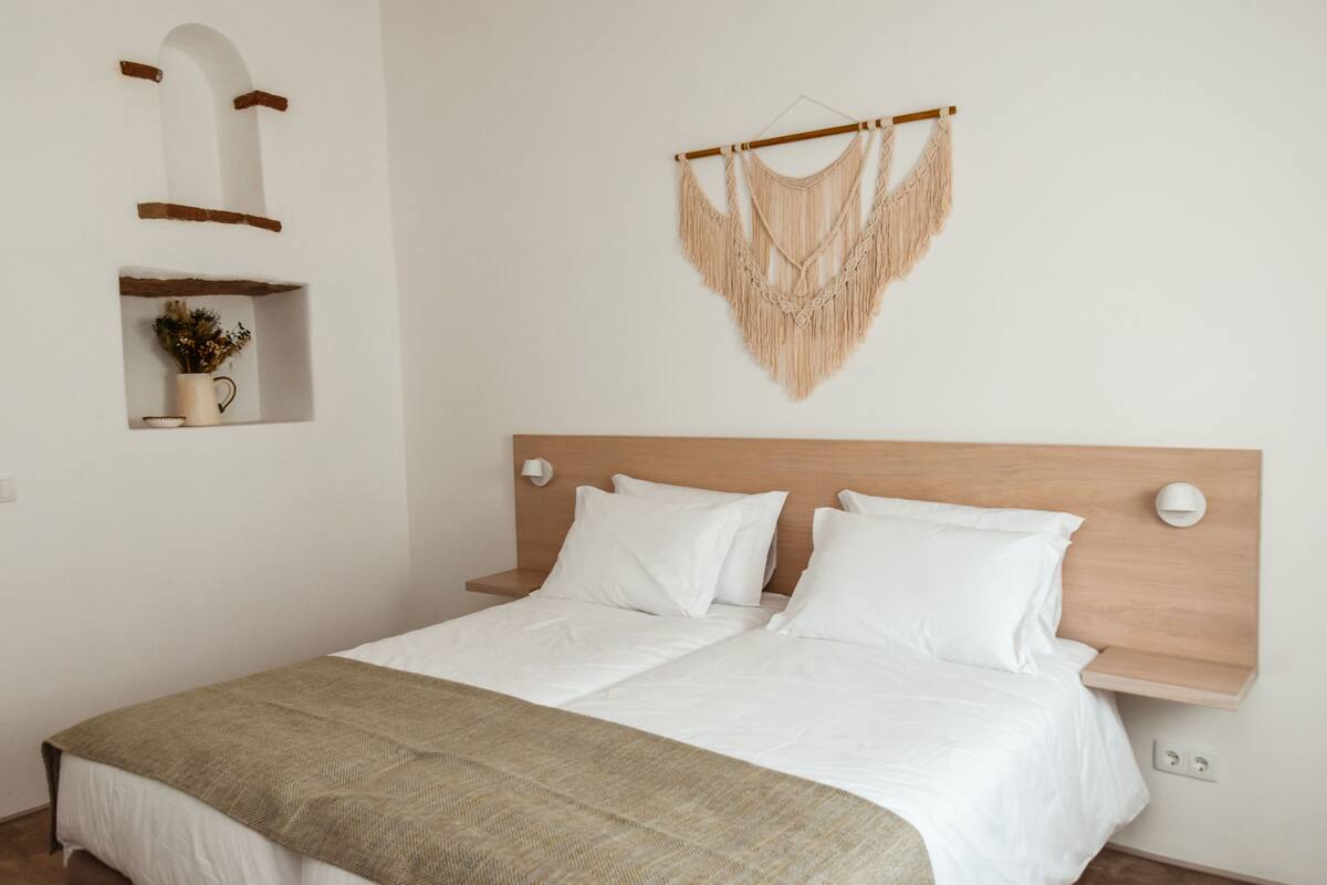 A neatly made bed with white bedding and a beige throw blanket, a wooden headboard, a wall-mounted macrame piece, and a shelf with a vase and flowers.